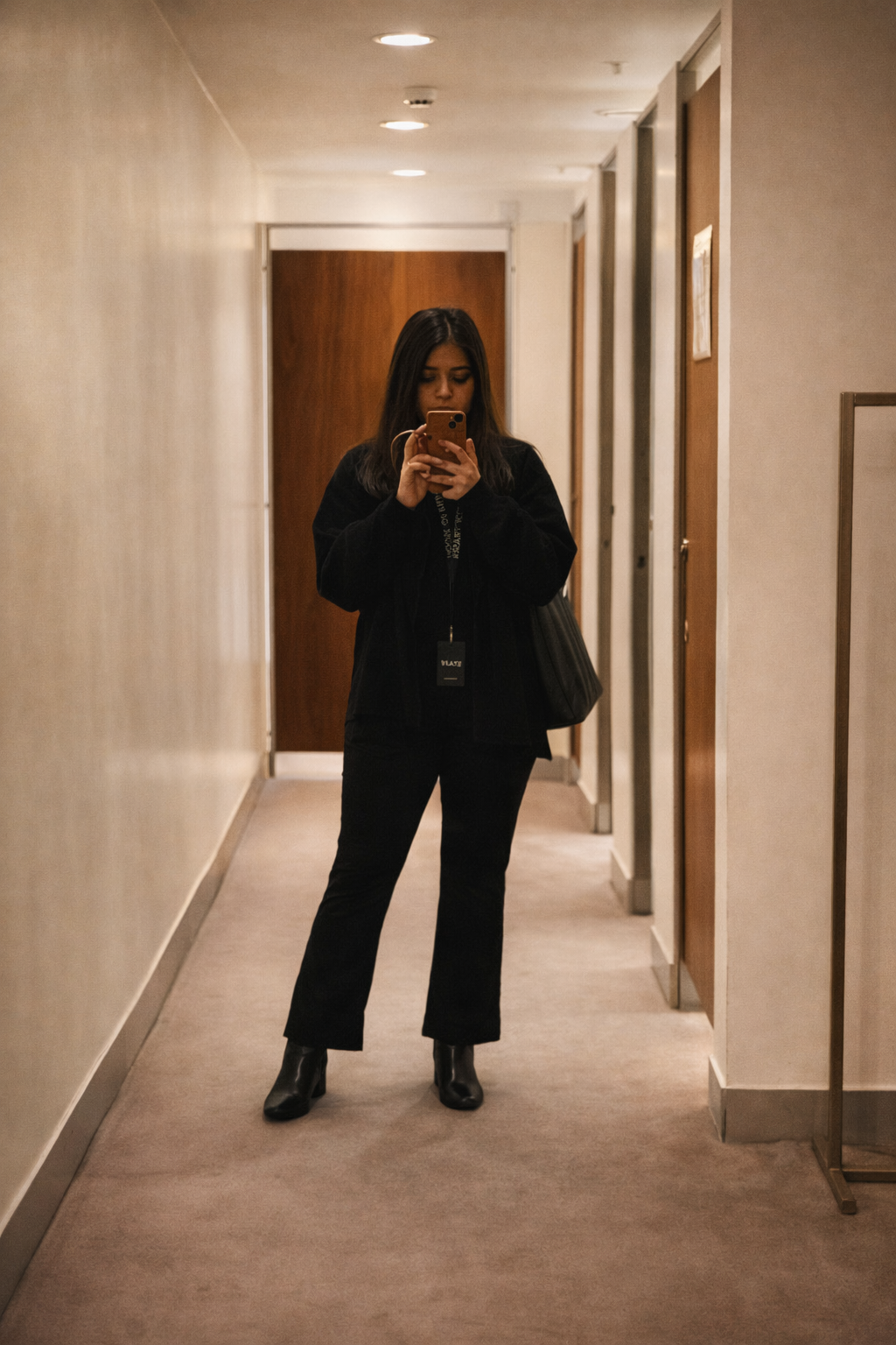 Woman taking a mirror selfie in a hallway outside fitting rooms, wearing an all-black outfit with ankle boots and a tote bag.