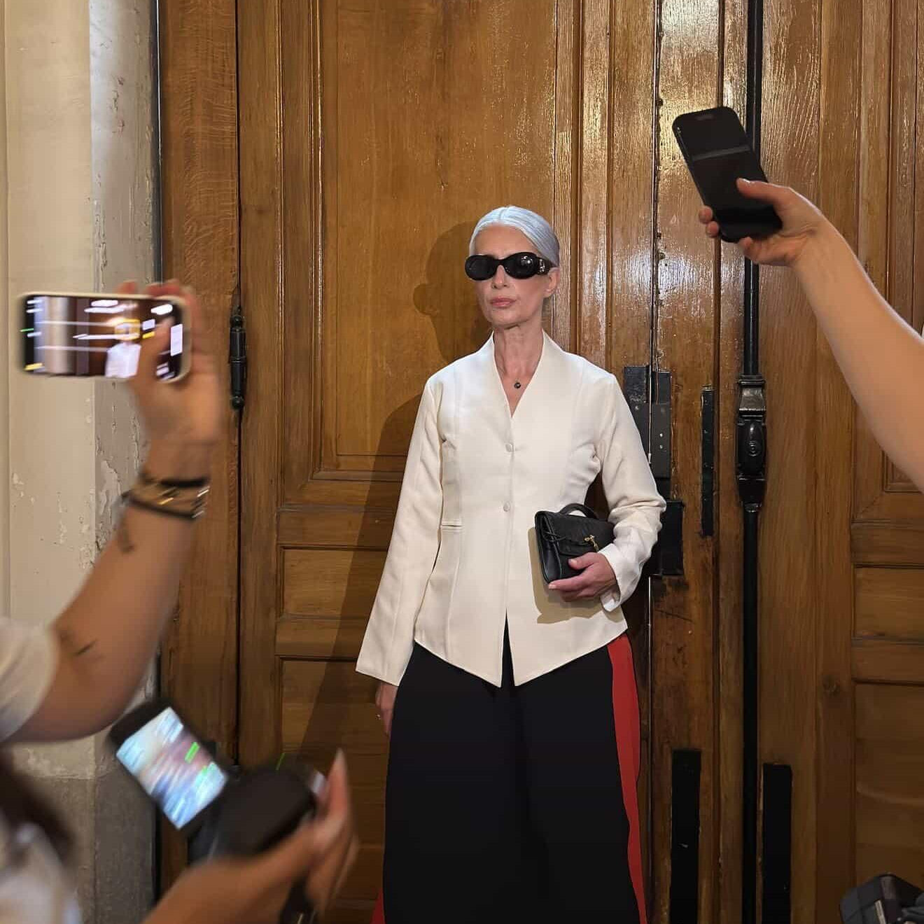 Person in a white shirt and black pants with red stripe standing in front of a wooden door with a circular window.